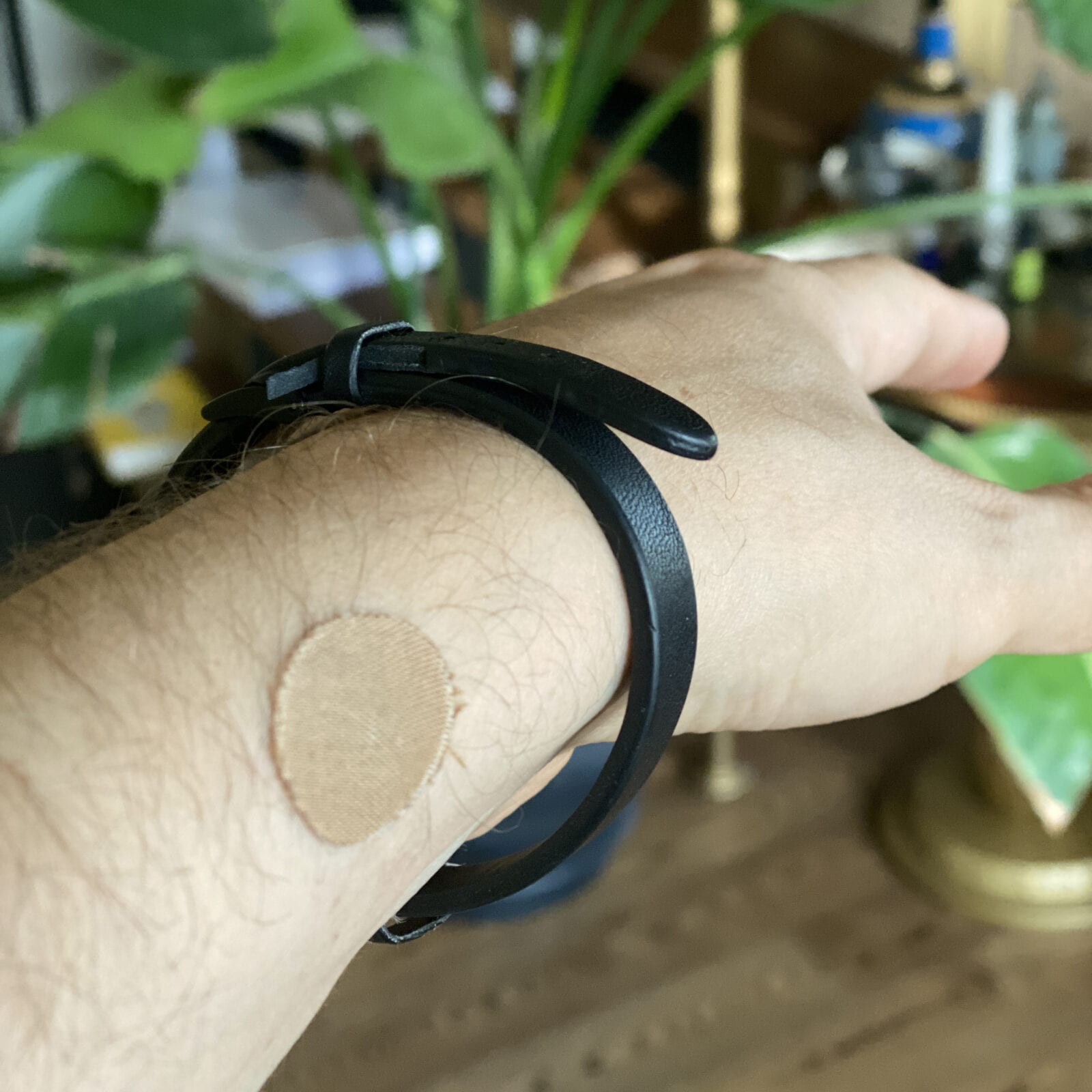 Forearm with a small beige adhesive patch below a black leather bracelet, plants blurred behind